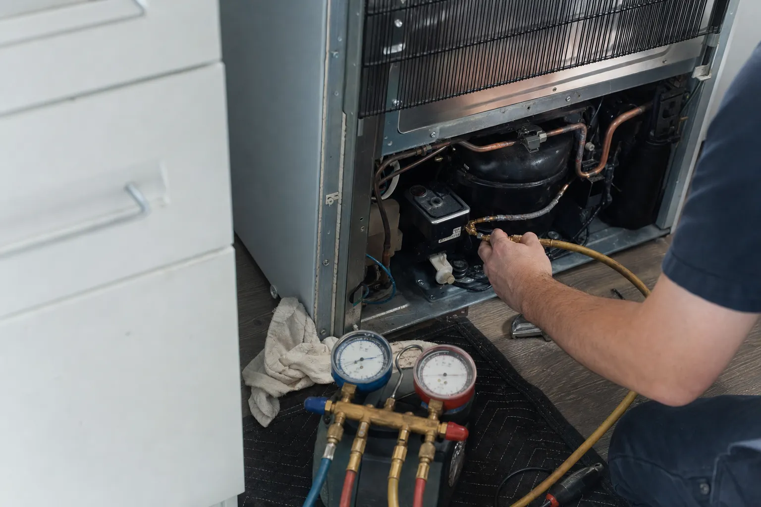 Refrigerator pulled clear of the cabinets with the rear machine compartment open, a charging hose fitted to the compressor