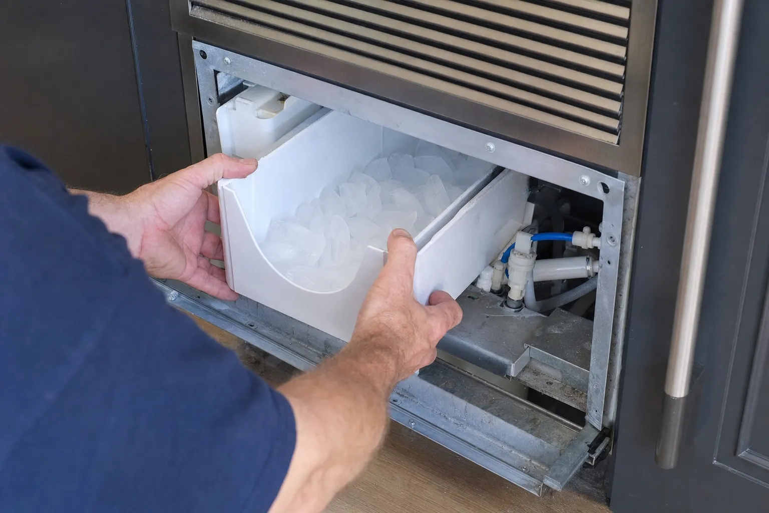 Hands pulling a white ice bin out of an undercounter ice maker beneath a louvred grille, blue water-line fittings beside it